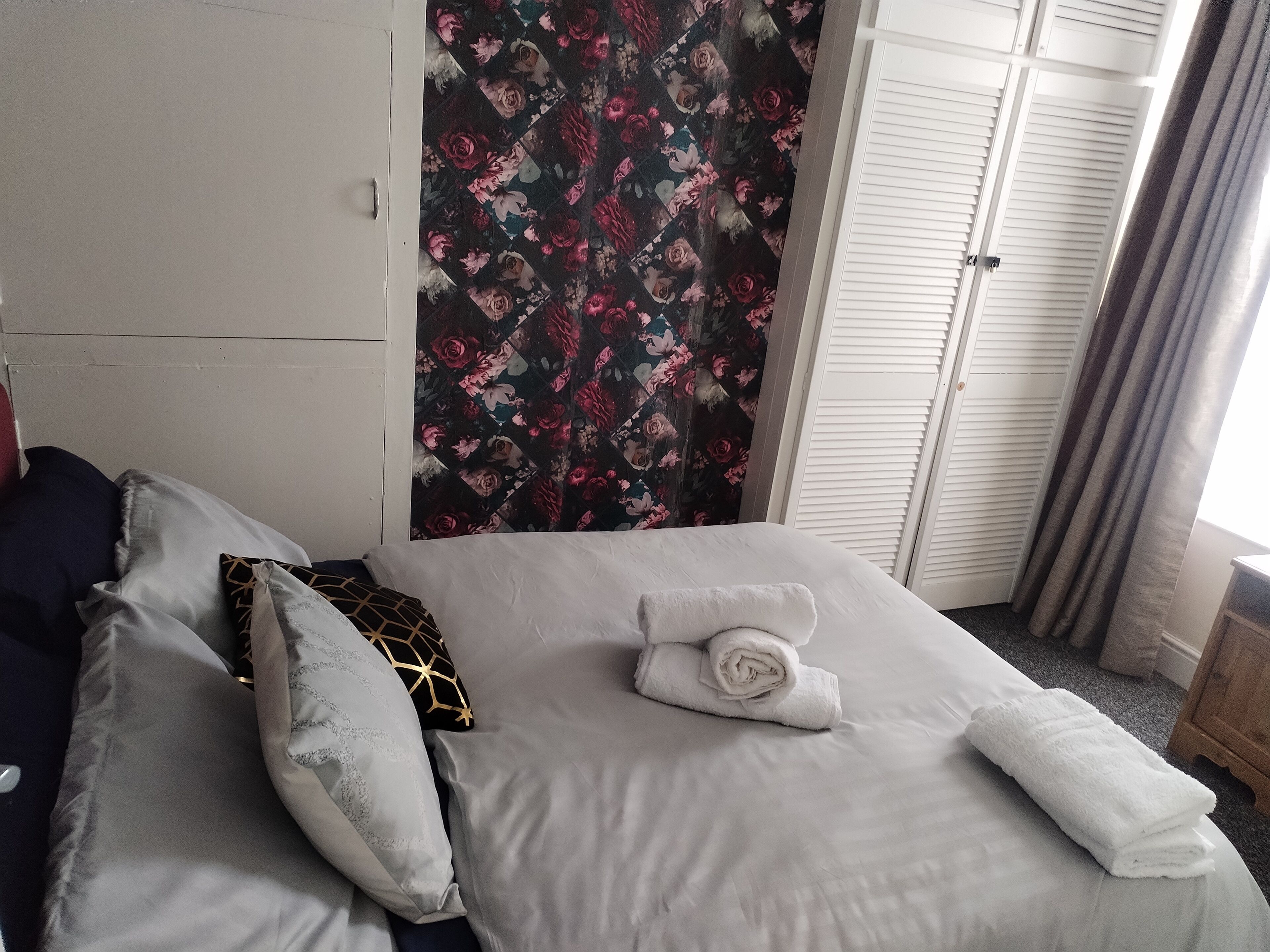 A neatly made bed with white linens and a rolled towel, a floral curtain, and a white wardrobe with louvered doors.