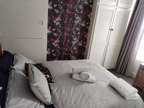 A neatly made bed with white linens and a rolled towel, a floral curtain, and a white wardrobe with louvered doors. - Comfortable 4-bed House in Hucknall, Nottingham (Nottingham)