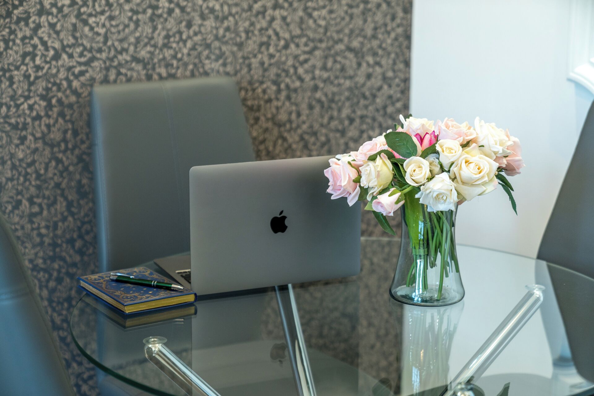 A laptop on a glass table with a vase of flowers.