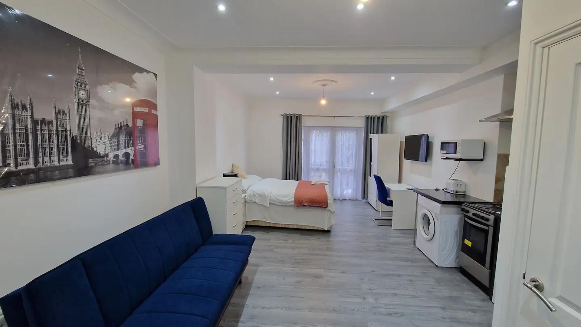Habitación de hotel con cama, sofá azul, televisor, lavadora y zona de cocina.