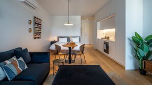 Een moderne, compacte woonruimte met een bank, eettafel, stoelen, een klein keukentje en een grote plant.
