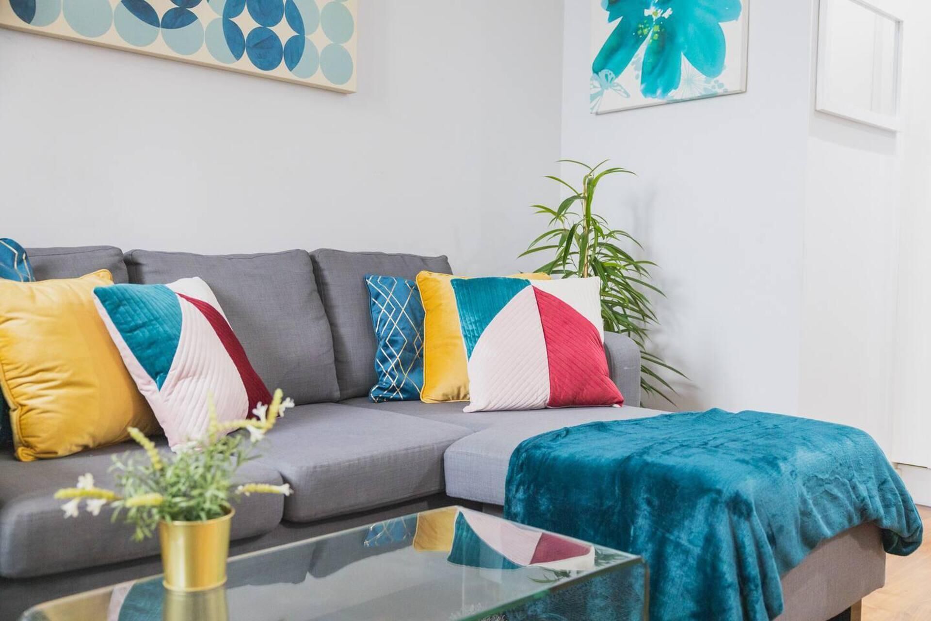 A modern living room with a grey sofa, colorful cushions, a glass coffee table, and a blue blanket.