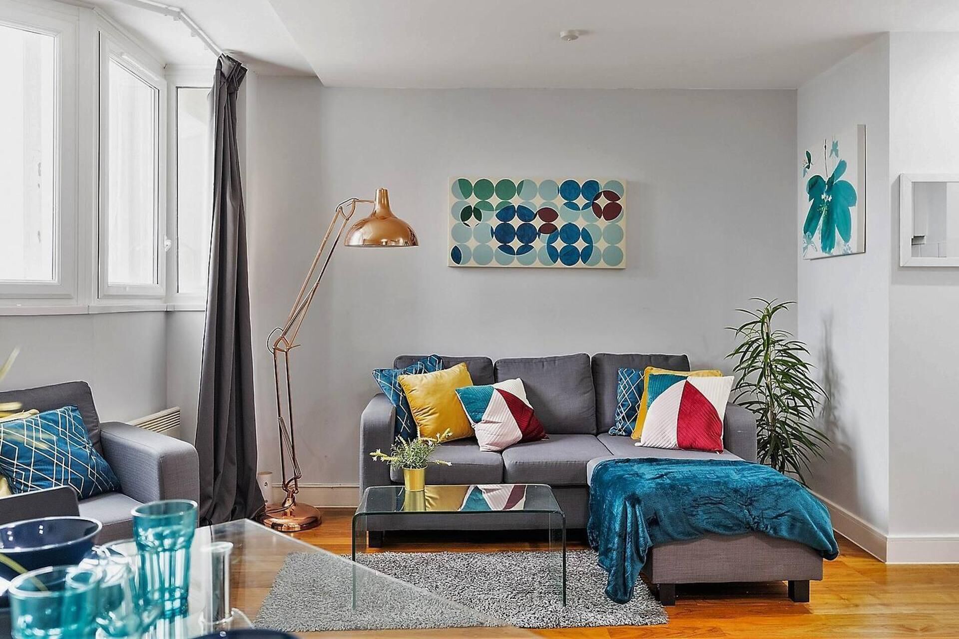 A modern living room with a grey sofa, colorful cushions, a floor lamp, and a geometric wall art piece.