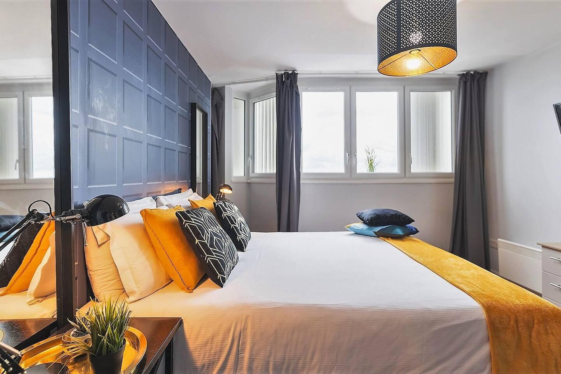 Moderni hotellihuone, jossa on suuri sänky, työpöytä lampun kanssa ja näkymä kaupunkiin suurista ikkunoista.