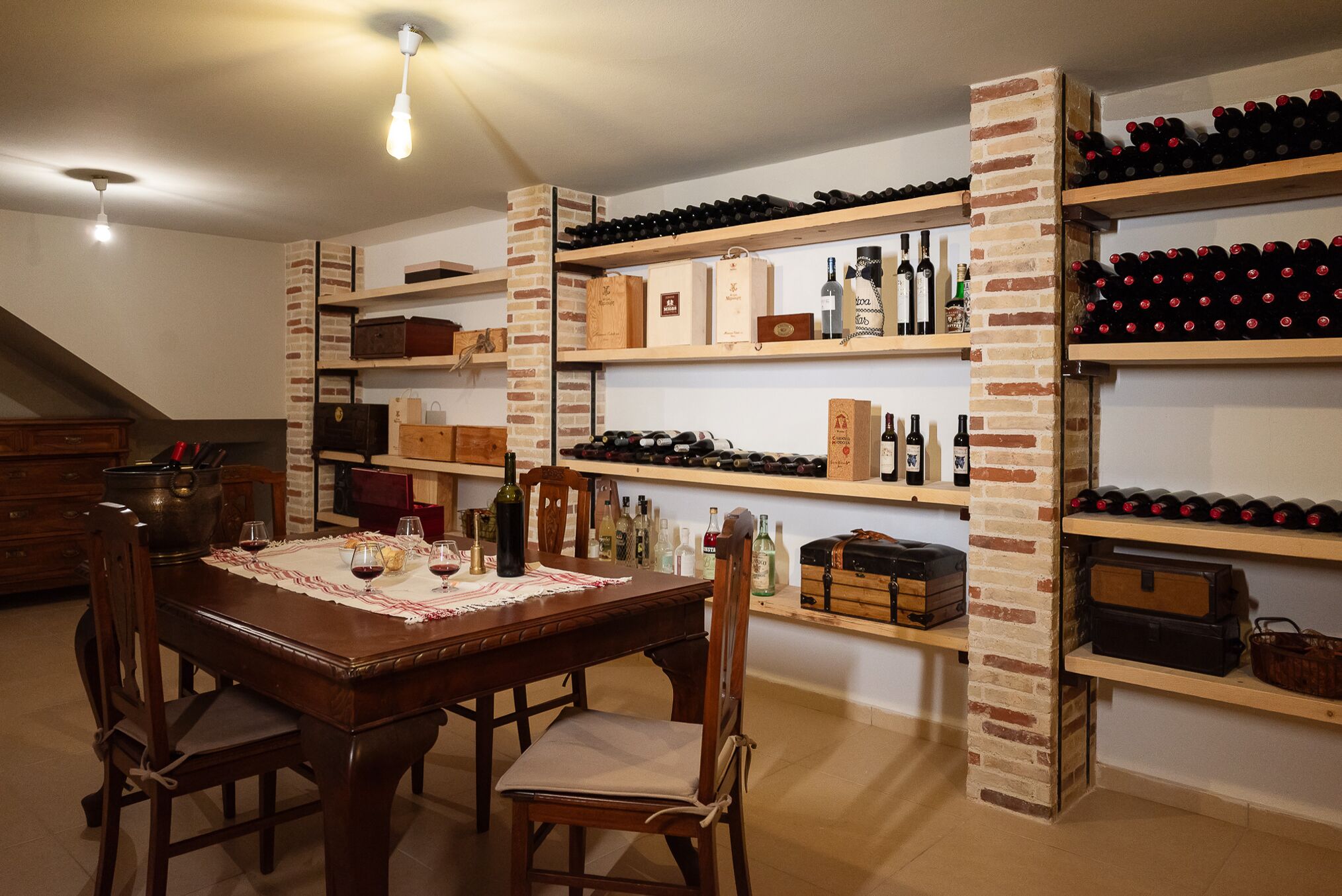 A dining area with a wooden table set for two, surrounded by wine bottles and boxes on brick shelving.