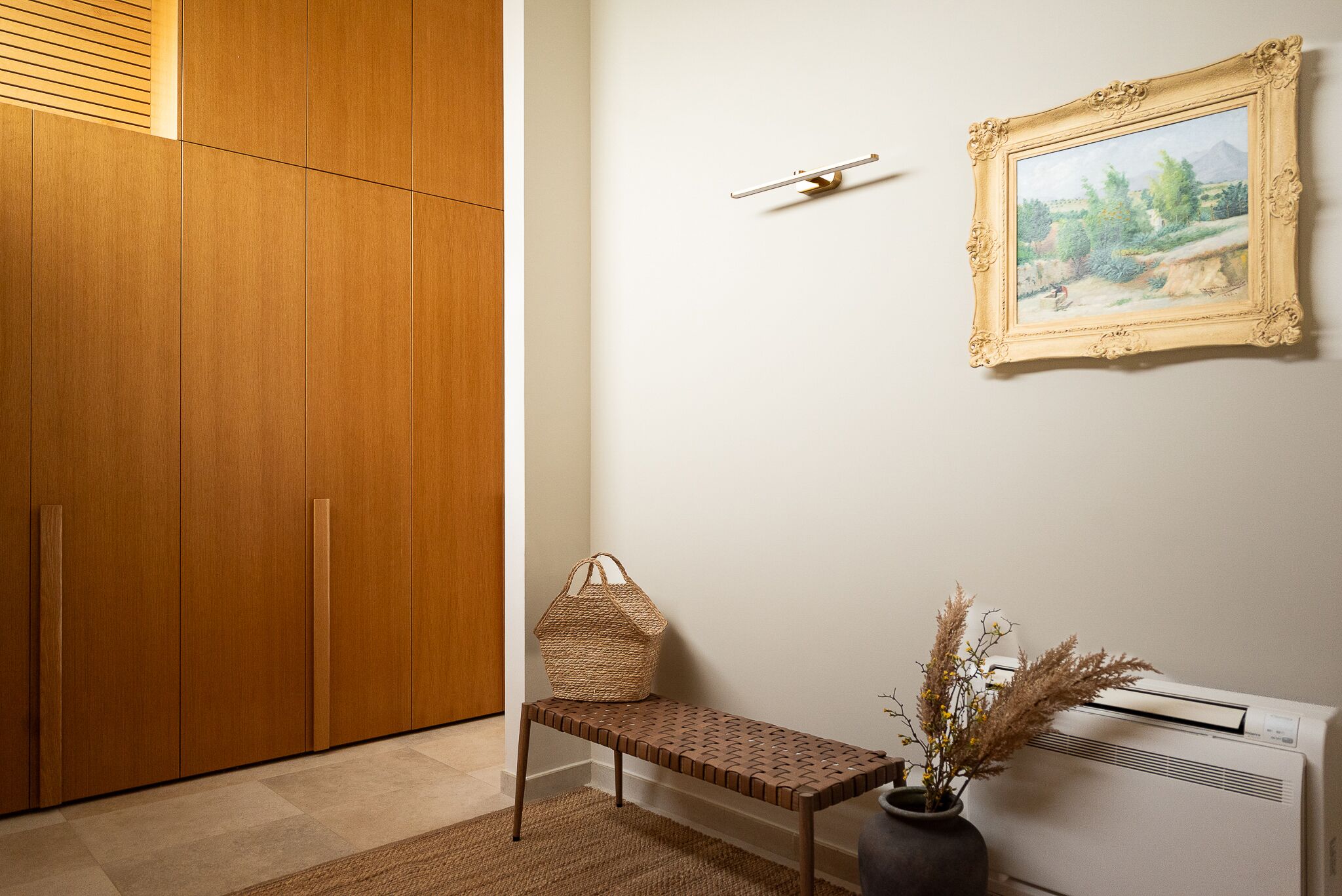 A room with a wooden wardrobe, a bench with a woven bag, a framed painting, and an air conditioning unit.
