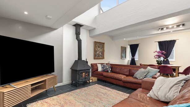 Een moderne woonkamer met een grote flatscreen televisie, een houten entertainmentmeubel, een bruine leren bank, een zwarte houtkachel, en een eethoek met tafel en stoelen.