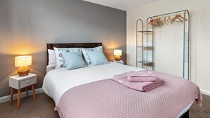 Een net opgemaakt bed met een roze deken, twee bedlampen en een metalen garderobe met kledinghangers.