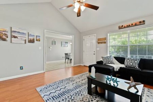 A spacious living room with a ceiling fan, a black sofa with patterned cushions, a coffee table with decorative items, and a large window with blinds.