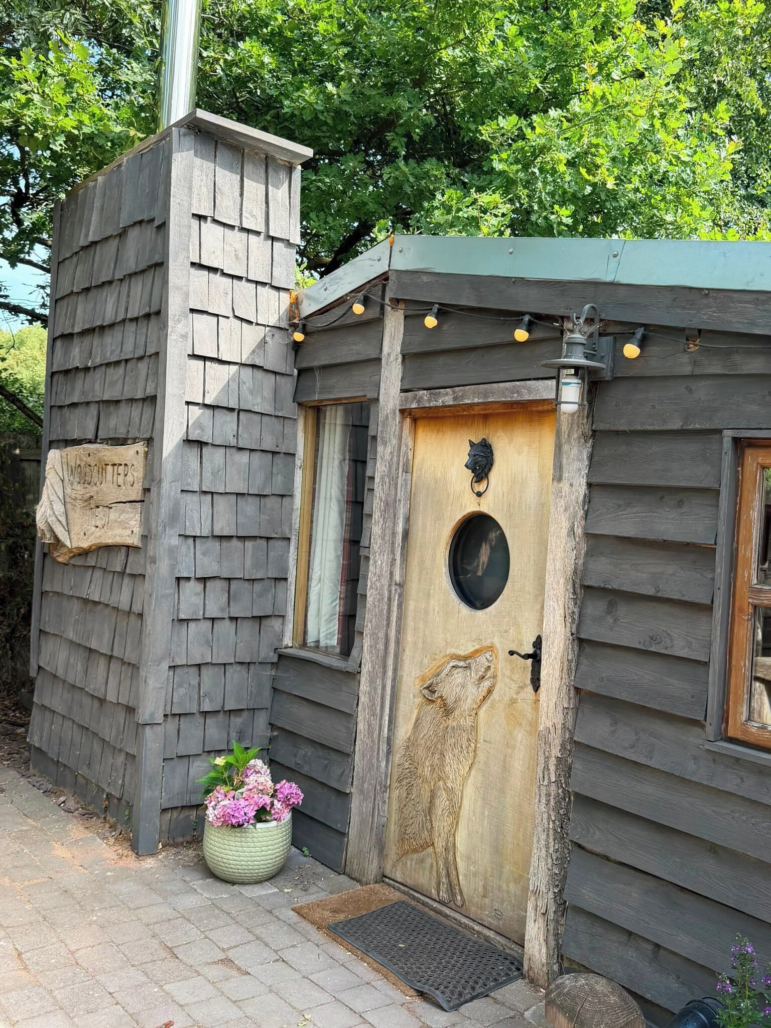 Une cabane rustique en bois, avec un revêtement extérieur en bardeaux, une porte en bois ornée d’une fenêtre circulaire et un petit porche agrémenté d’une plante en pot.