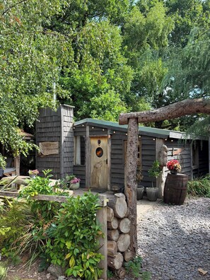 A rustic cabin with a wooden door, surrounded by greenery and a gravel path. - The Woodcutters Rest Cabin Sutton Coldfield (Sutton Coldfield)