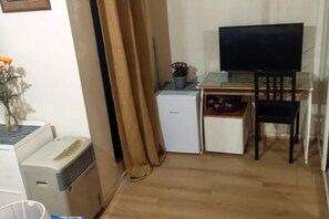 Une chambre de taille modeste, équipée d’un bureau, d’un petit réfrigérateur, d’une chaise et d’un téléviseur.