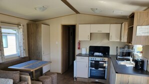 A compact kitchen with a stove, oven, and sink, a dining area with a table and chairs, and a small seating area with cushions. - Static Caravan - The Dizzard (Holsworthy)