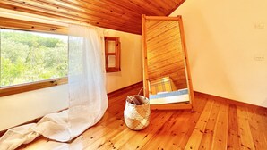 A room with wooden flooring, a window with a view of greenery, a small wooden bed frame, and a woven basket on the floor.