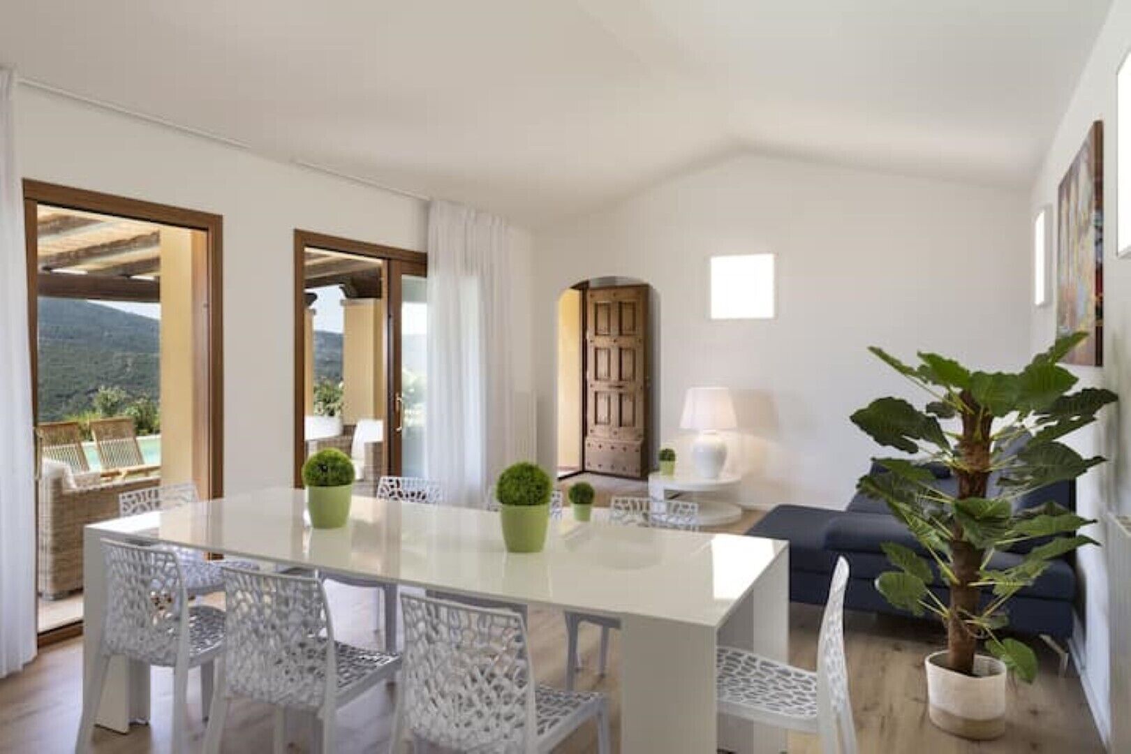 A spacious living area with a large dining table, white chairs, a blue sofa, and a potted plant.