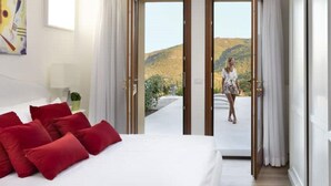A hotel room with a large bed, red pillows, a painting on the wall, and a view of a hilly landscape through open doors.
