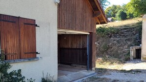 A rustic wooden building with a garage door open, gravel driveway, and a stone wall in the background.