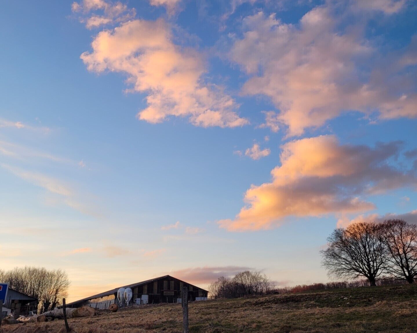 Un paysage rural avec une grange en bois, de vastes champs ouverts et un ciel dégagé parsemé de quelques nuages.