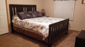 A bedroom with a bed, a bedside table, a lamp, a window with blinds, and a wall-mounted fan. - Home Away from Home (Natchez)