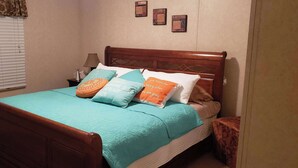A neatly made bed with a teal bedspread and multiple pillows, a wooden headboard, a bedside table with a lamp, and a window with blinds. - Home Away from Home (Natchez)