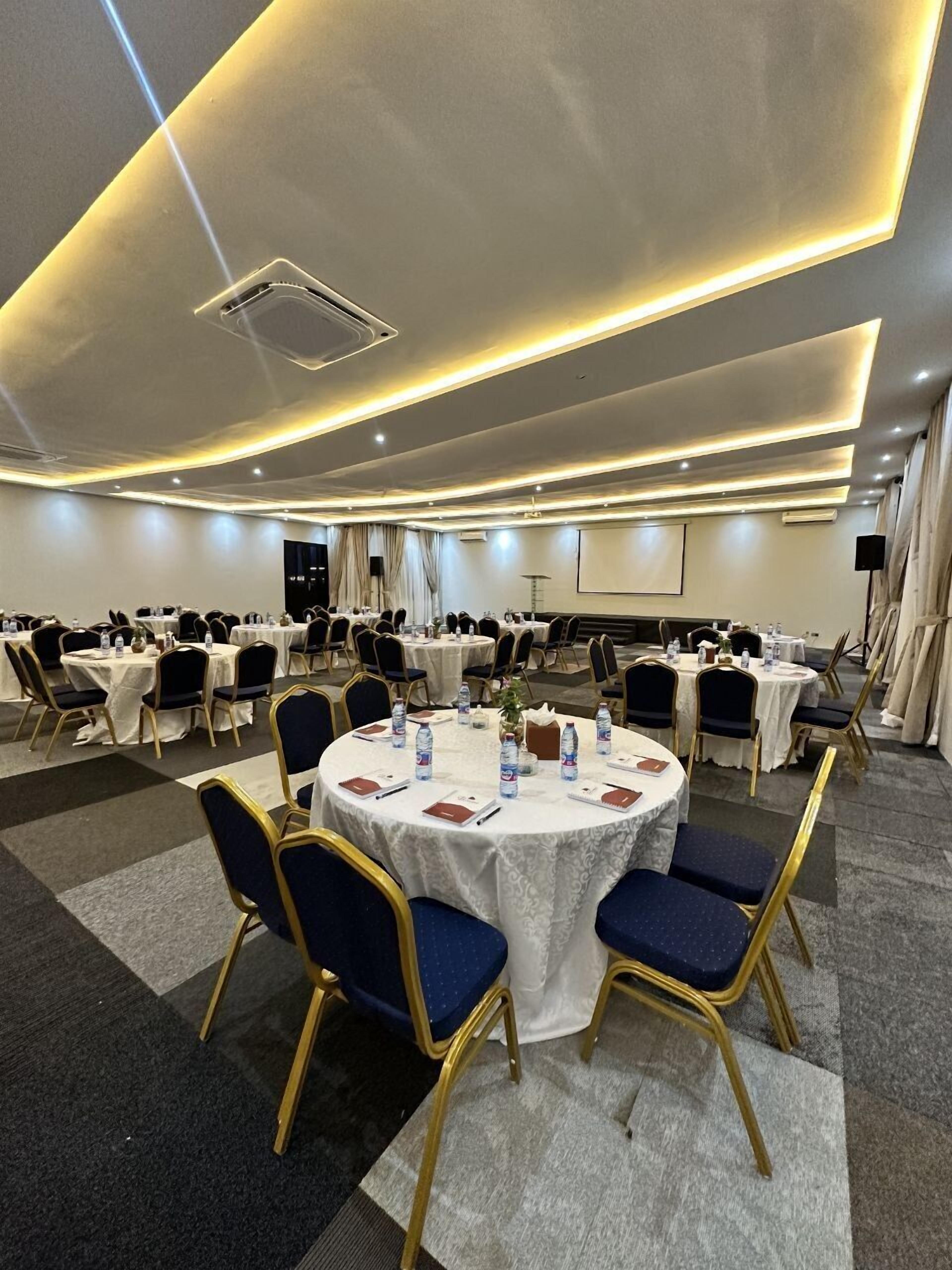 Un salón de conferencias con mesas redondas, sillas y una pantalla para proyector.