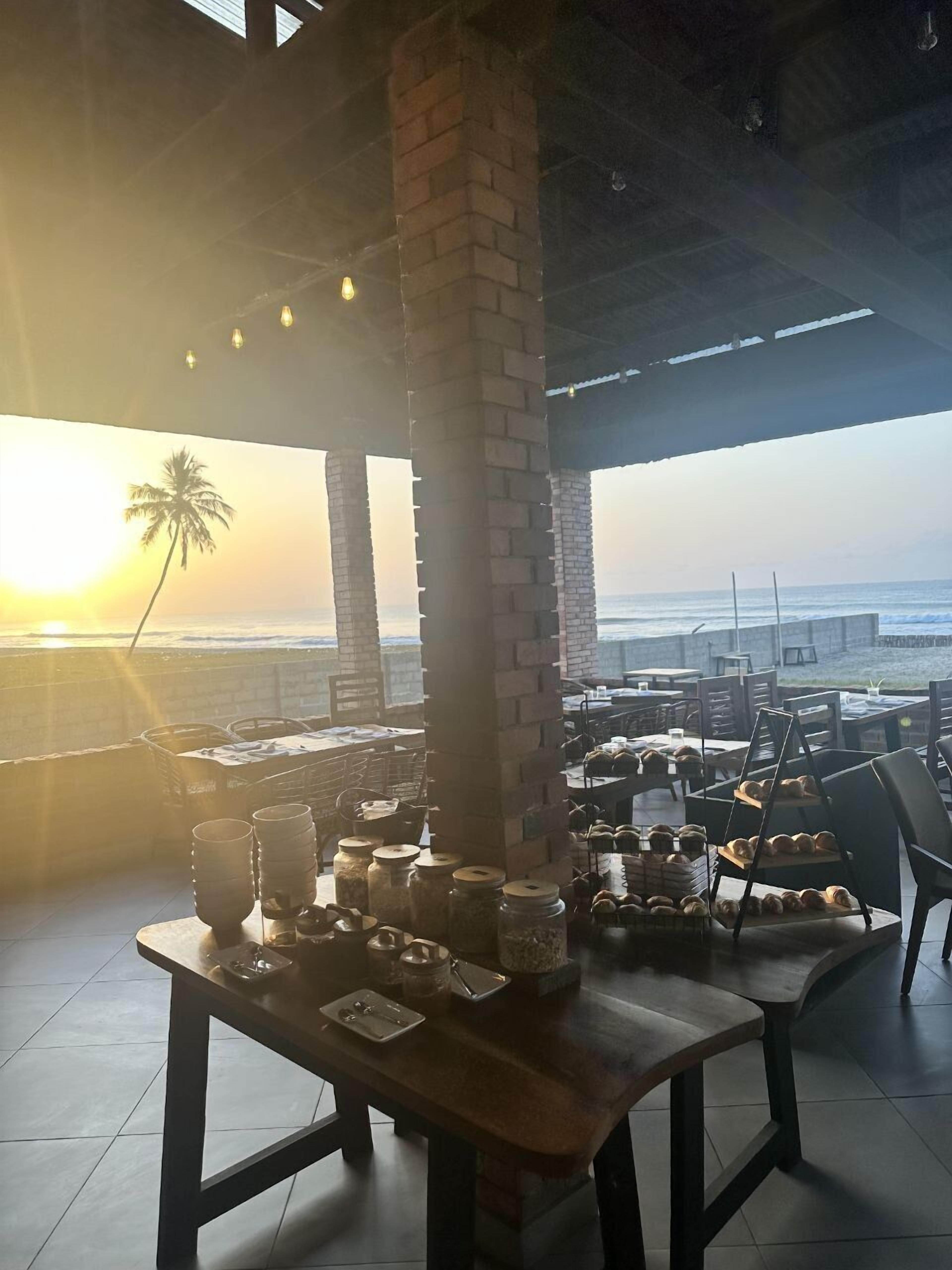 A beachfront restaurant with exposed brick pillars, outdoor seating, and a view of the ocean at sunset.