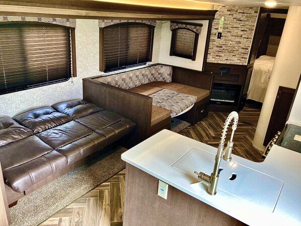 Interior de una caravana compacta con sofá, una pequeña cocina y una ventana con persianas.