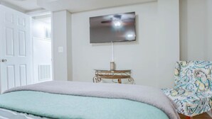 A bedroom with a bed, a TV mounted on the wall, a small table with a lamp, and a patterned chair. - Cozy Basement Suite with Full Kitchen, Walk to Edgewood (Atlanta)