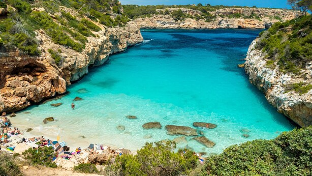 A coastal area with clear turquoise waters, rocky cliffs, and a beach with people sunbathing.