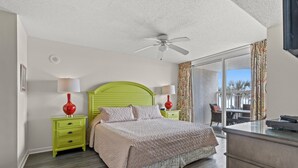 A bedroom with a green headboard, a bed with a beige quilt, two bedside tables with lamps, a ceiling fan, and a view of a balcony with outdoor furniture. - Updated Crescent Shores 107 - Oceanfront 3/3 Condo (North Myrtle Beach)