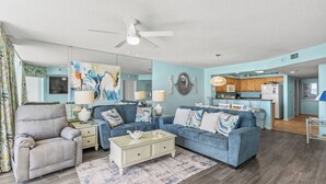 A living room with a sofa, armchair, and coffee table, a kitchen in the background, and a ceiling fan. - Updated Crescent Shores 107 - Oceanfront 3/3 Condo (North Myrtle Beach)