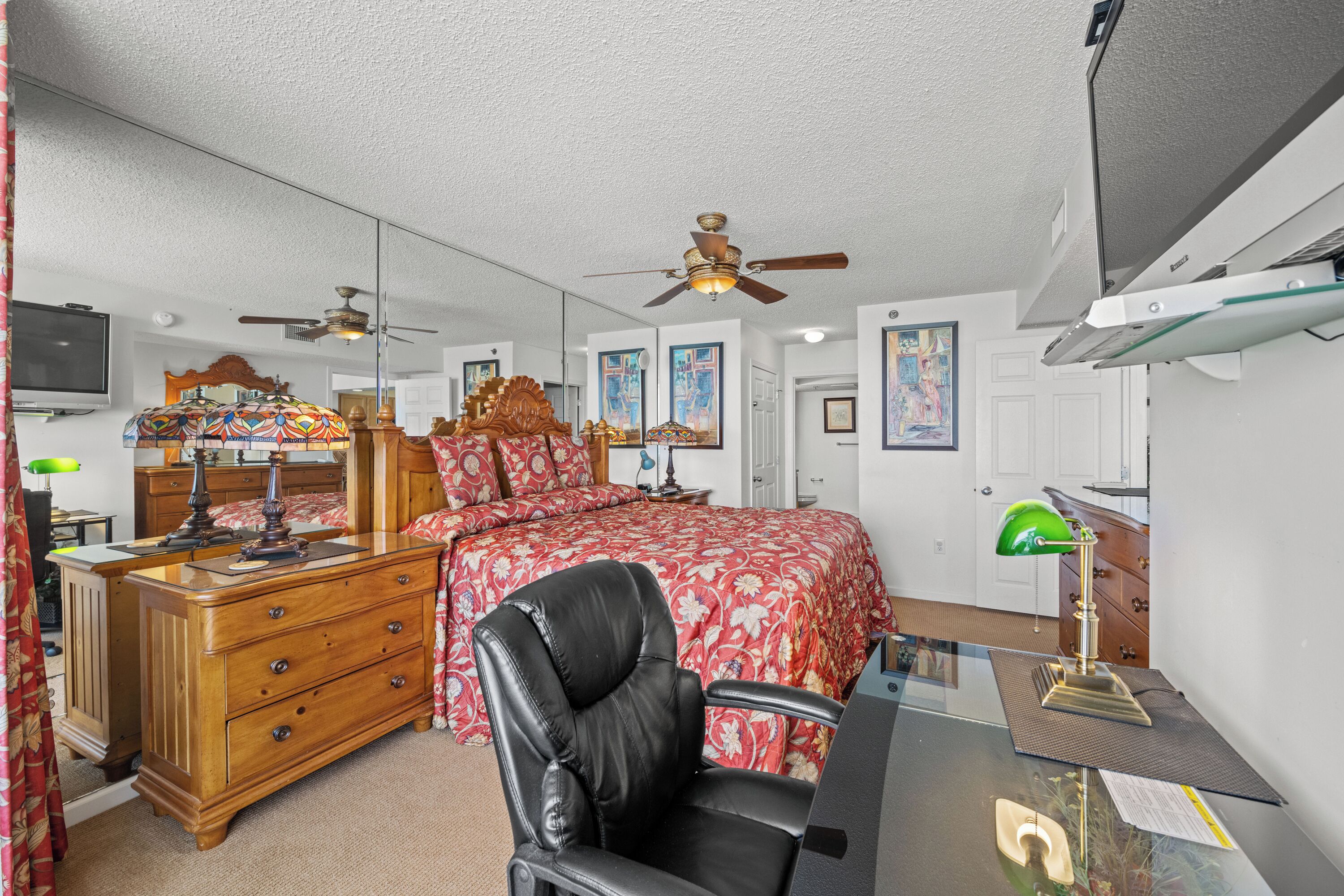 A bedroom with a bed, a desk, a chair, a dresser, a mirror, and a ceiling fan.