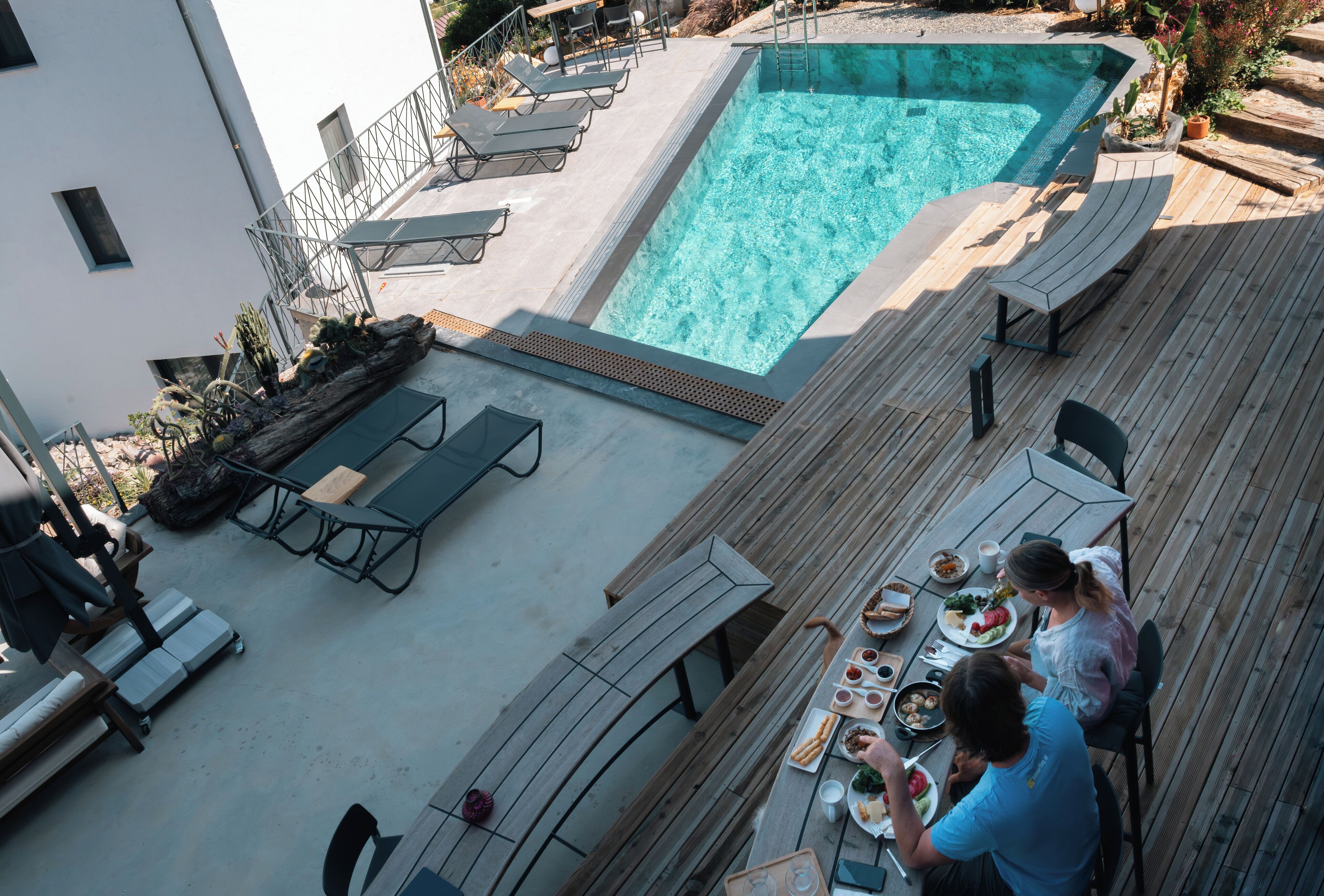 A rooftop pool area with a dining table and chairs, surrounded by lounge chairs and a wooden deck.