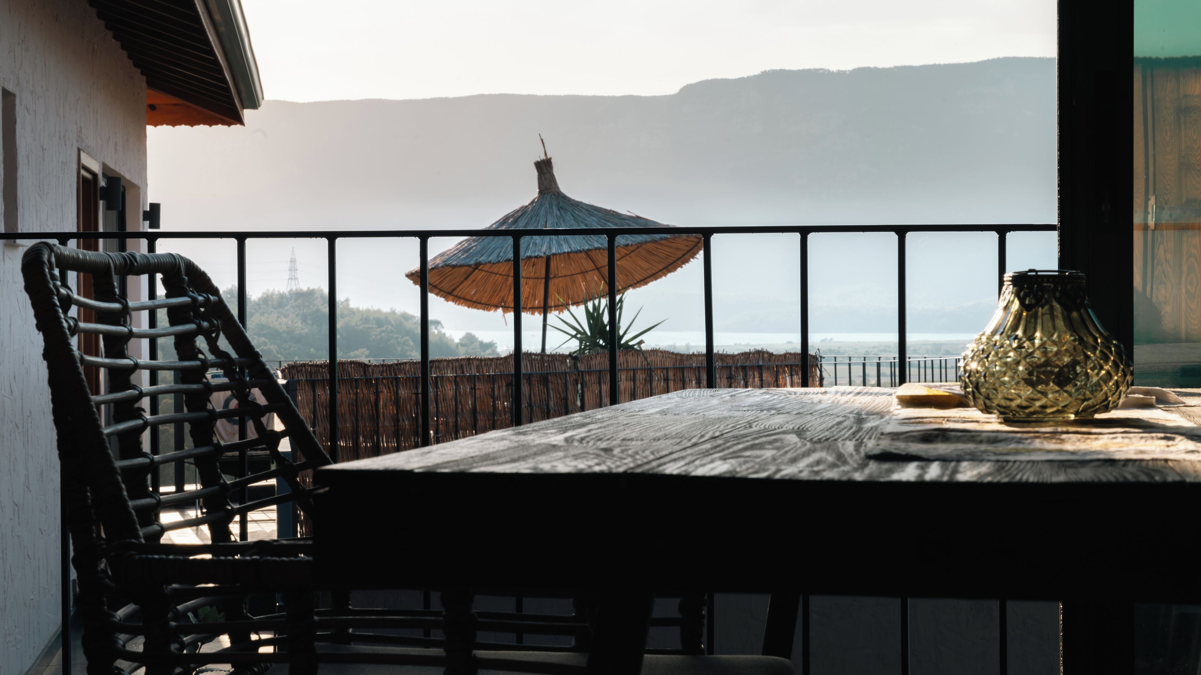 A balcony with a table, a wicker chair, a glass vase, and a straw umbrella.