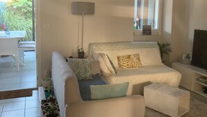 Een moderne woonkamer met een bank, voetenbankje en een tv-meubel. Er is een raam met uitzicht op de natuur, een lamp en een potplant.
