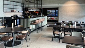 Un restaurante moderno con techo alto, vigas de madera a la vista y pared de ladrillo. Hay varias mesas y sillas, una zona de bar y un área de estar con vistas al exterior.