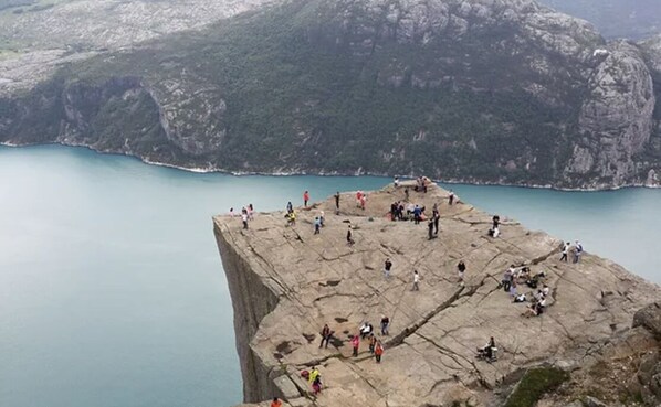 Un acantilado rocoso con gente reunida en el borde, contemplando una masa de agua tranquila y un paisaje montañoso.