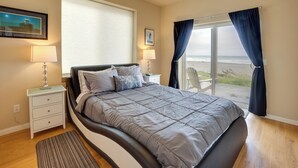 A bedroom with a bed, nightstands, a window with curtains, and a view of the beach. - Gold Beach Retreat: Private Hot Tub + Ocean Views! (Gold Beach)