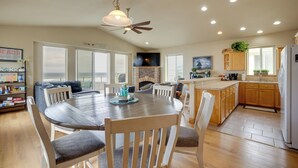 A spacious living area with a dining table, chairs, a fireplace, and a kitchen with wooden cabinets and a refrigerator. - Gold Beach Retreat: Private Hot Tub + Ocean Views! (Gold Beach)