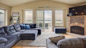 A living room with a large sectional sofa, a fireplace, and a view of the beach through glass doors. - Gold Beach Retreat: Private Hot Tub + Ocean Views! (Gold Beach)
