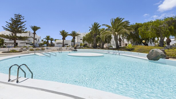 A swimming pool with a circular center, surrounded by palm trees and white buildings.