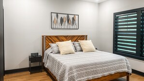 A bedroom with a wooden headboard, a bed with patterned bedding, a nightstand, a wall art piece, and a window with blinds.