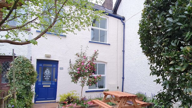A white house with a blue door, a small garden with flowers and a wooden table. - Bay Tree Cottage (Keswick)