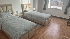 Een slaapkamer met twee bedden, houten hoofdeinden, een raam met gordijnen en uitzicht op gebouwen.