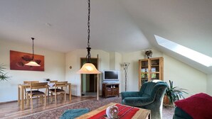 Ein geräumiges Wohnzimmer mit hoher Decke, einer Essecke mit Holzmöbeln, einem Fernseher und einem großen Fenster, das für natürliches Licht sorgt.