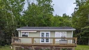 Une cabane en bois à deux étages, dotée d’une terrasse et entourée d’une pelouse et d’arbres.