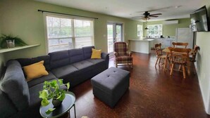 Un salon comprenant un canapé, un pouf et une petite table sur laquelle est posée une plante. On y trouve également un coin repas avec des chaises en bois et une table. La pièce est dotée d’un ventilateur de plafond et, au fond, on aperçoit un coin cuisine.