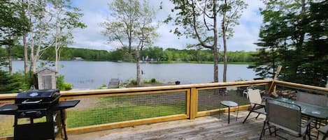 A wooden deck with a grill, chairs, and a table overlooking a lake.