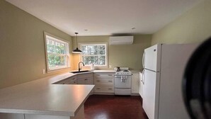 Une cuisine avec des meubles blancs, une cuisinière et un réfrigérateur. Il y a des fenêtres qui donnent sur un paysage verdoyant.