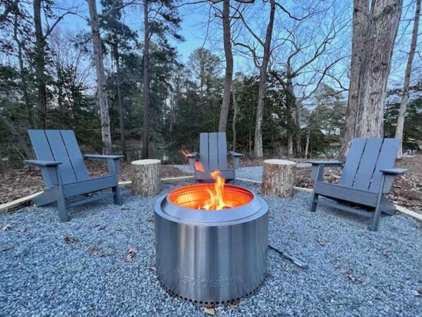 Una fogata metálica circular con llama visible, rodeada por tres sillas Adirondack y dos troncos de árbol.